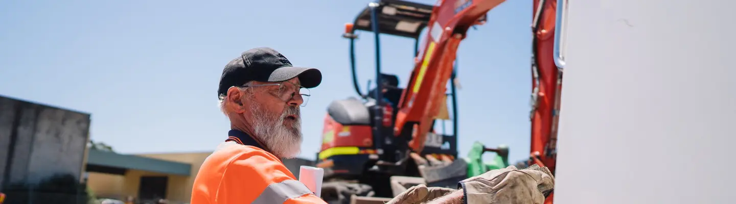 Worker standing beside a loader truck at a construction site, inspecting or adjusting equipment near safety barriers.