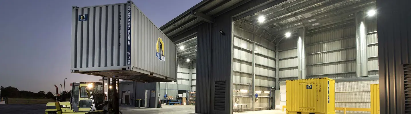 Forklift lifting a shipping container outside a large industrial warehouse with illuminated interior and additional containers inside.