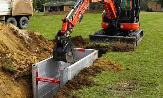 Worker operating excavation machinery near braced trench with visible hazardous material signs on a construction site.