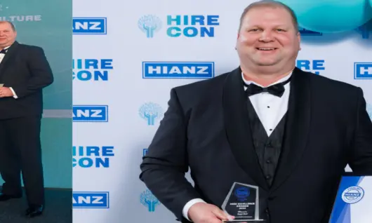 Two employees smiling and holding awards, standing in front of a company logo backdrop at a recognition event.