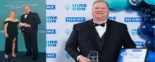 Two employees smiling and holding awards, standing in front of a company logo backdrop at a recognition event.