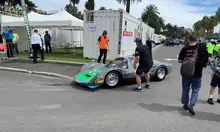 Workers setting up shipping containers and parked race cars at a festival site prepared for a motorsport event.