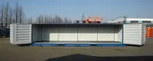 A large shipping container with its side fully opened, revealing a clean, empty interior space. The container is set outdoors in an industrial yard, with trees and equipment visible in the background, and appears suitable for storage or display use.